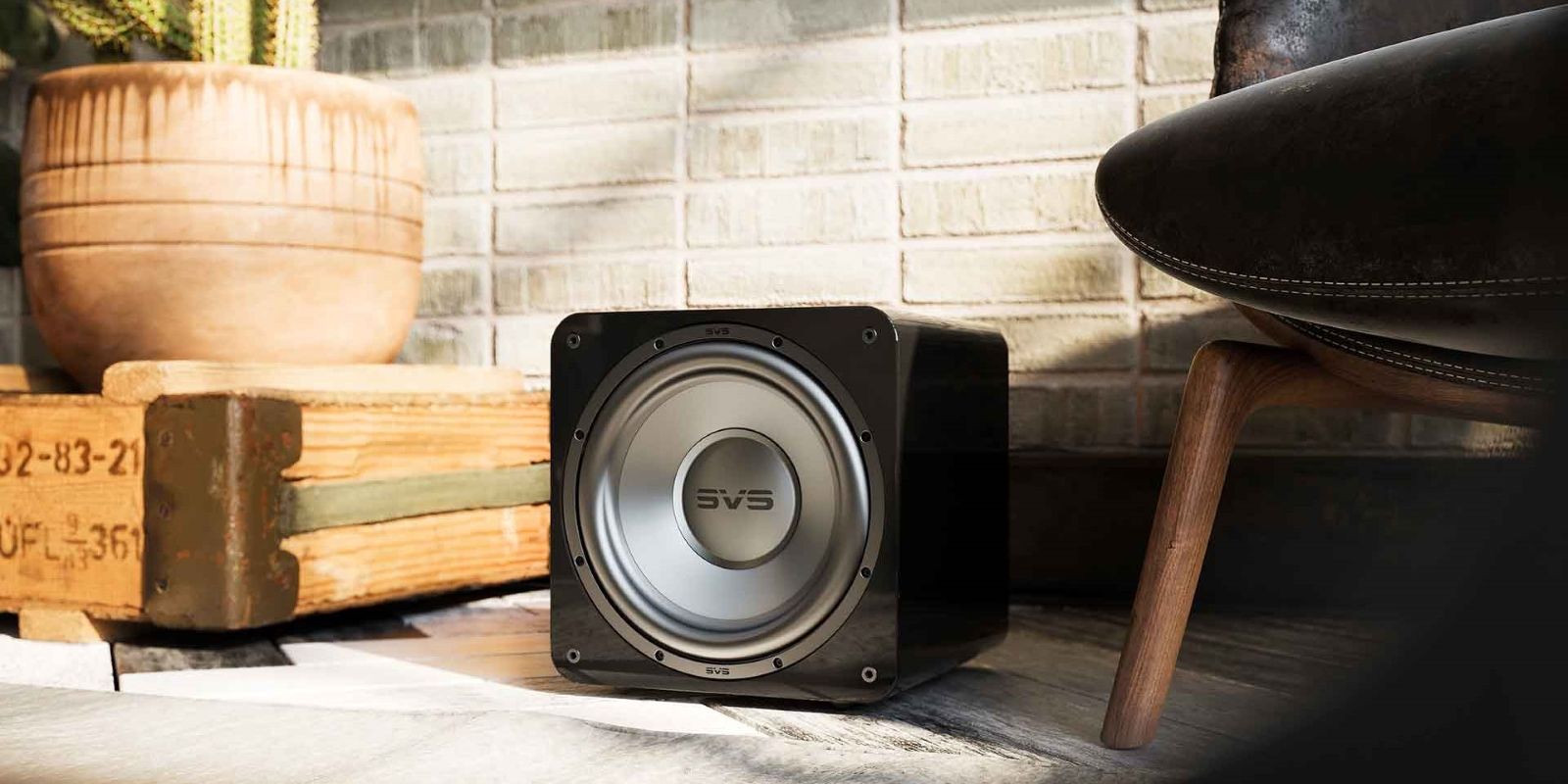 SVS SB1000 Pro Subwoofer displayed in a modern room, featuring a sleek, compact cube design with a silver front driver. It sits on a wooden surface, blending seamlessly with the stylish décor, including a potted plant and contemporary furniture.